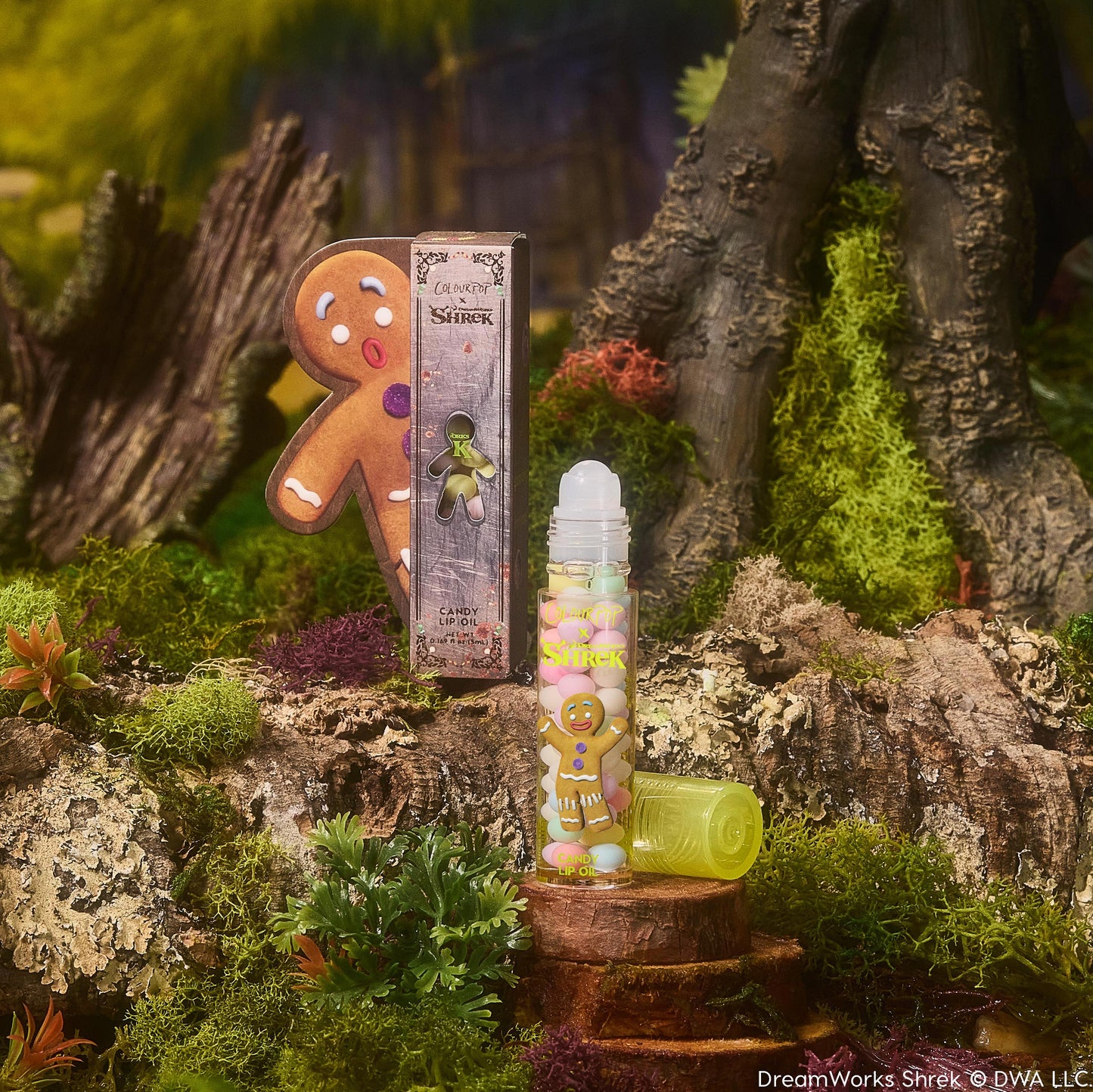 Gumdrop Buttons ColourPop Candy Lip Oil stands uncapped on a mossy stump, clear roll-on tube with gingerbread man graphic and lime cap beside it, posed with Shrek-themed packaging in a swampy forest setting.