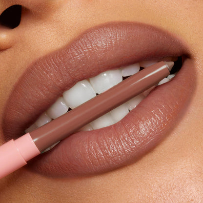 ColourPop So Juicy Plumping Lip Liner with Peptides in Tease defining full lips as the nude-brown pencil, with a blush-pink cap, is held between teeth, showcasing precise, creamy payoff and smooth contour in a studio close-up.